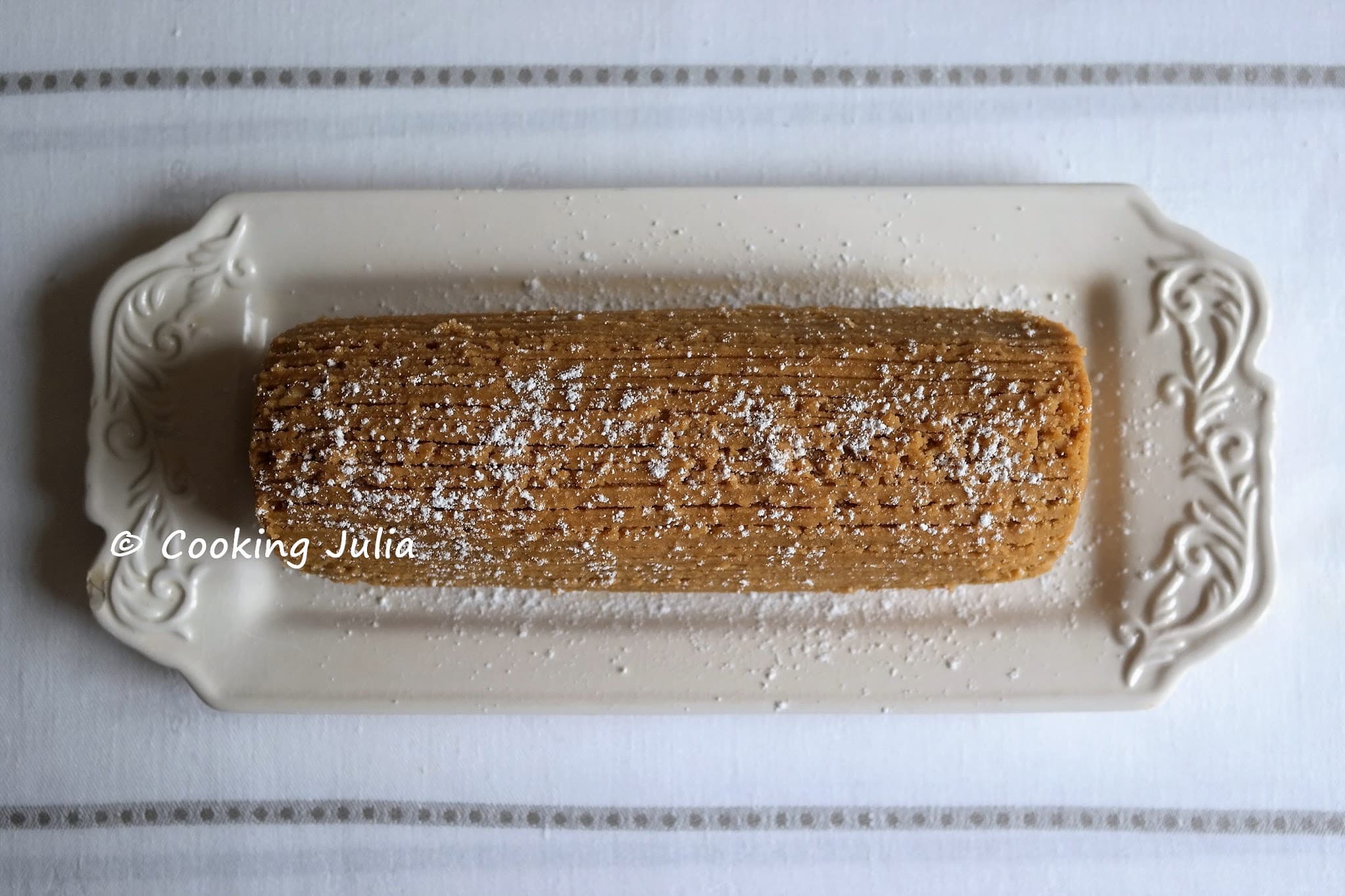 Bûche de Noël au café entière posée au centre d’un plat en porcelaine blanche décorée, saupoudrée de sucre glace